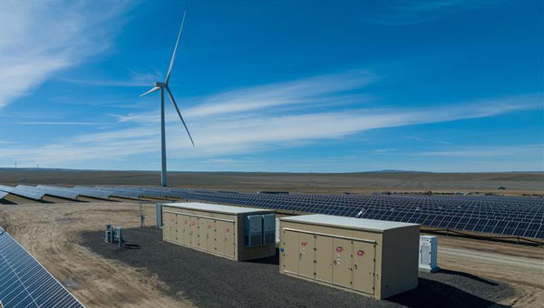 Two units at a battery storage facility surrounded by solar panels and a turbine in the distance
