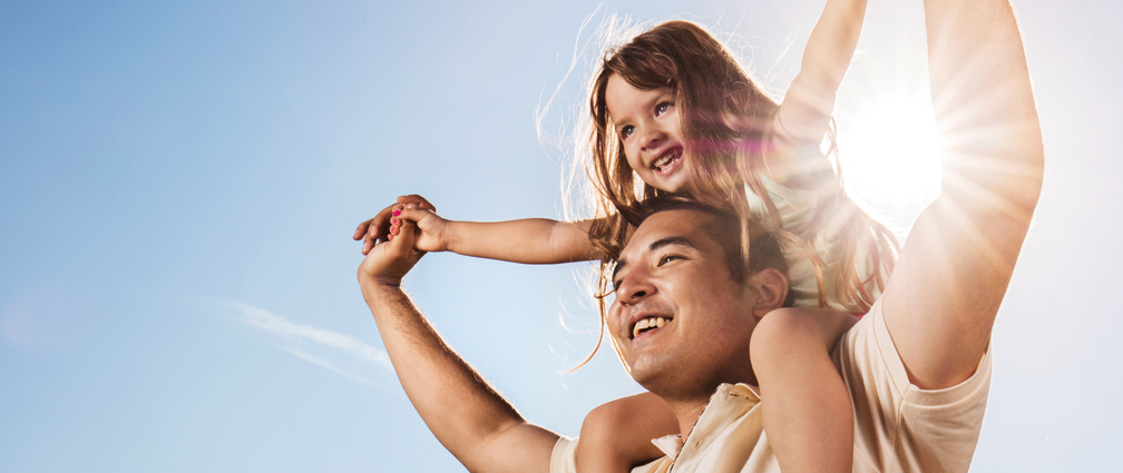 father with his younger kid daughter sitting on his shoulders outdoors under bright sunshine