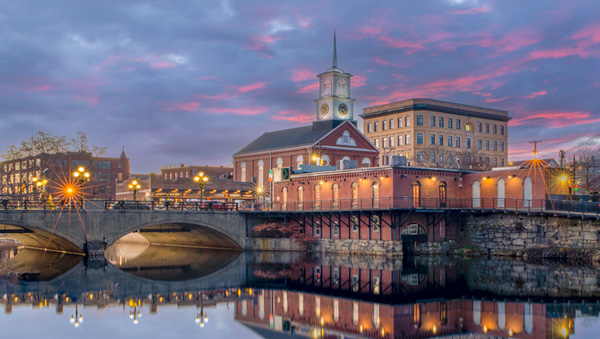 New England waterfront city at dusk with illuminated buildings and reflections on the water, representing reliable energy for the region.